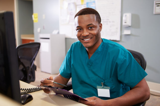 Portrait Of Male Nurse Working At Nurses Station Smiling At Camera
