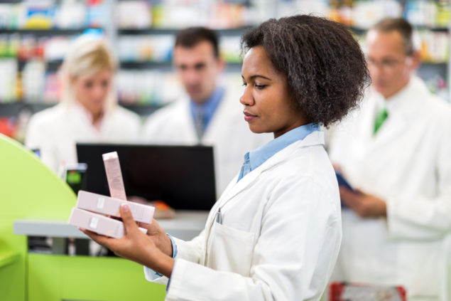 Group of pharmacists working in a pharmacy. Focus is on foreground, on a African American female pharmacist holding boxes of medicine.