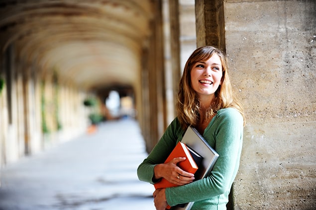 A female student stands in a hallway. She is leaning against the wall and her arms are filled with books.